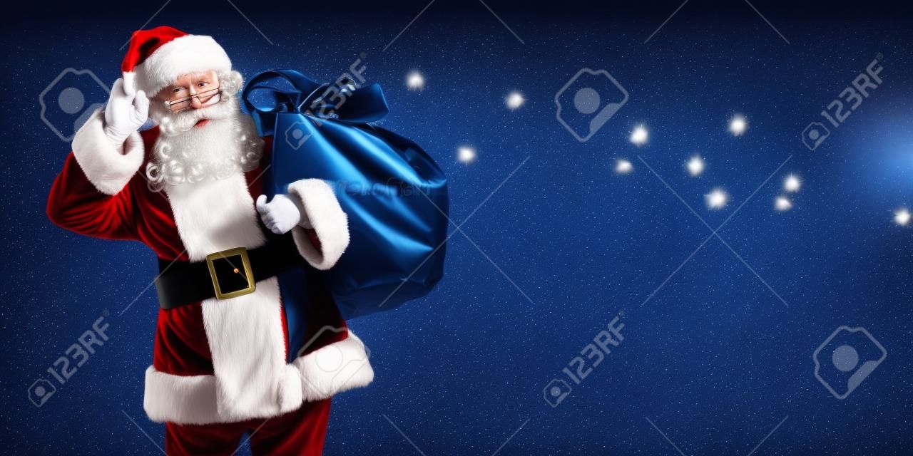 Der gute alte Weihnachtsmann steht mit einer Tüte voller Geschenke da und hört zu. Dunkelblauer Hintergrund mit leuchtenden Sternen. Weihnachten und Neujahr. Platz kopieren.