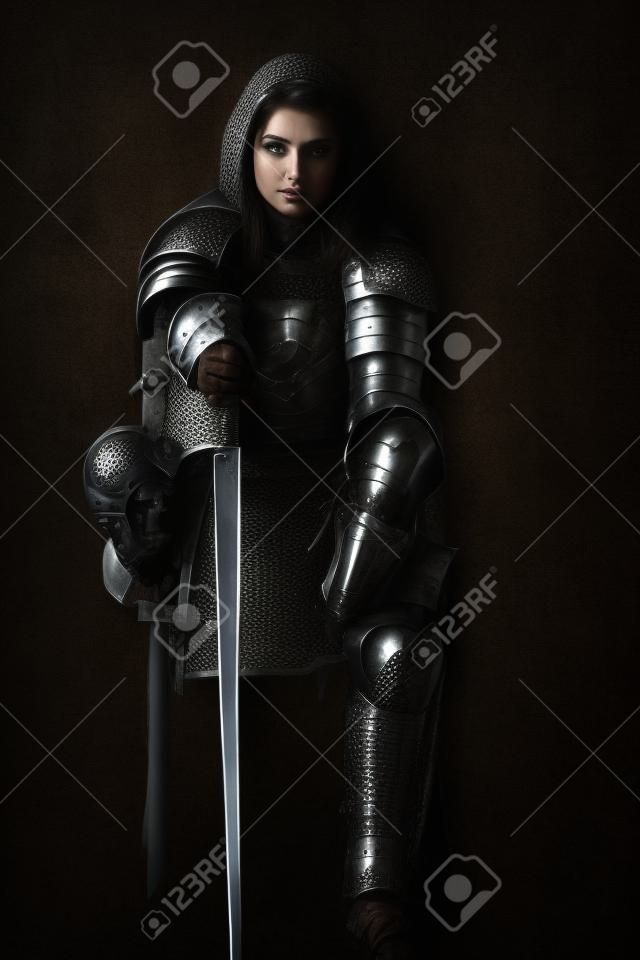 A beautiful noble warrior woman in chain mail and plate armor sits, leaning on her sword. Medieval knight. Studio portrait on a black background.