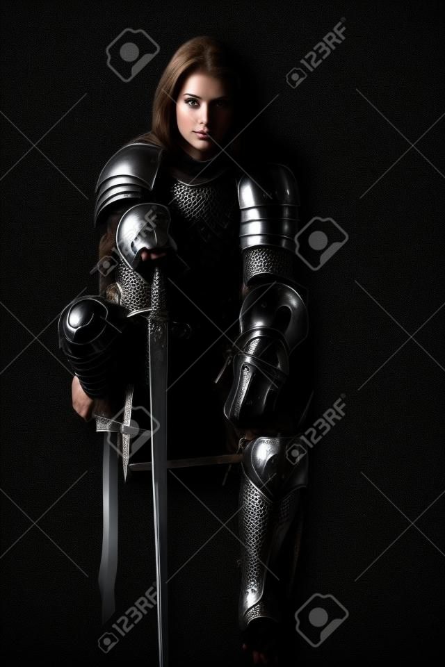 Piękna szlachetna wojowniczka w kolczudze i zbroi płytowej siedzi, opierając się na swoim mieczu. średniowieczny rycerz. portret studyjny na czarnym tle.