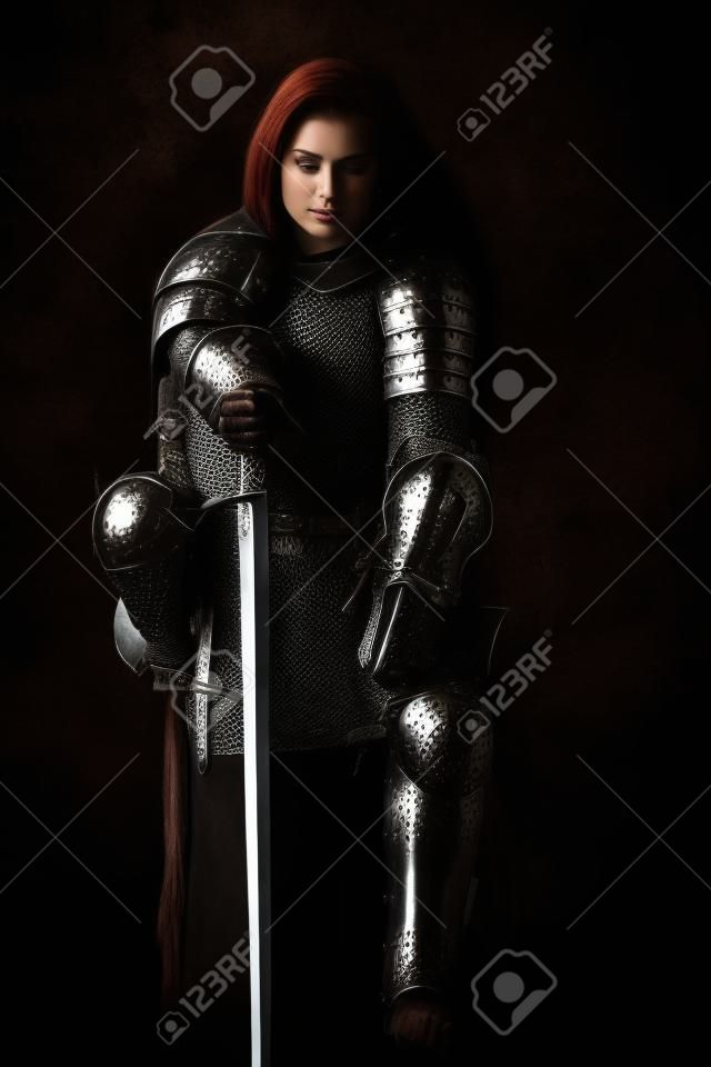 A beautiful noble warrior woman in chain mail and plate armor sits, leaning on her sword. Medieval knight. Studio portrait on a black background.