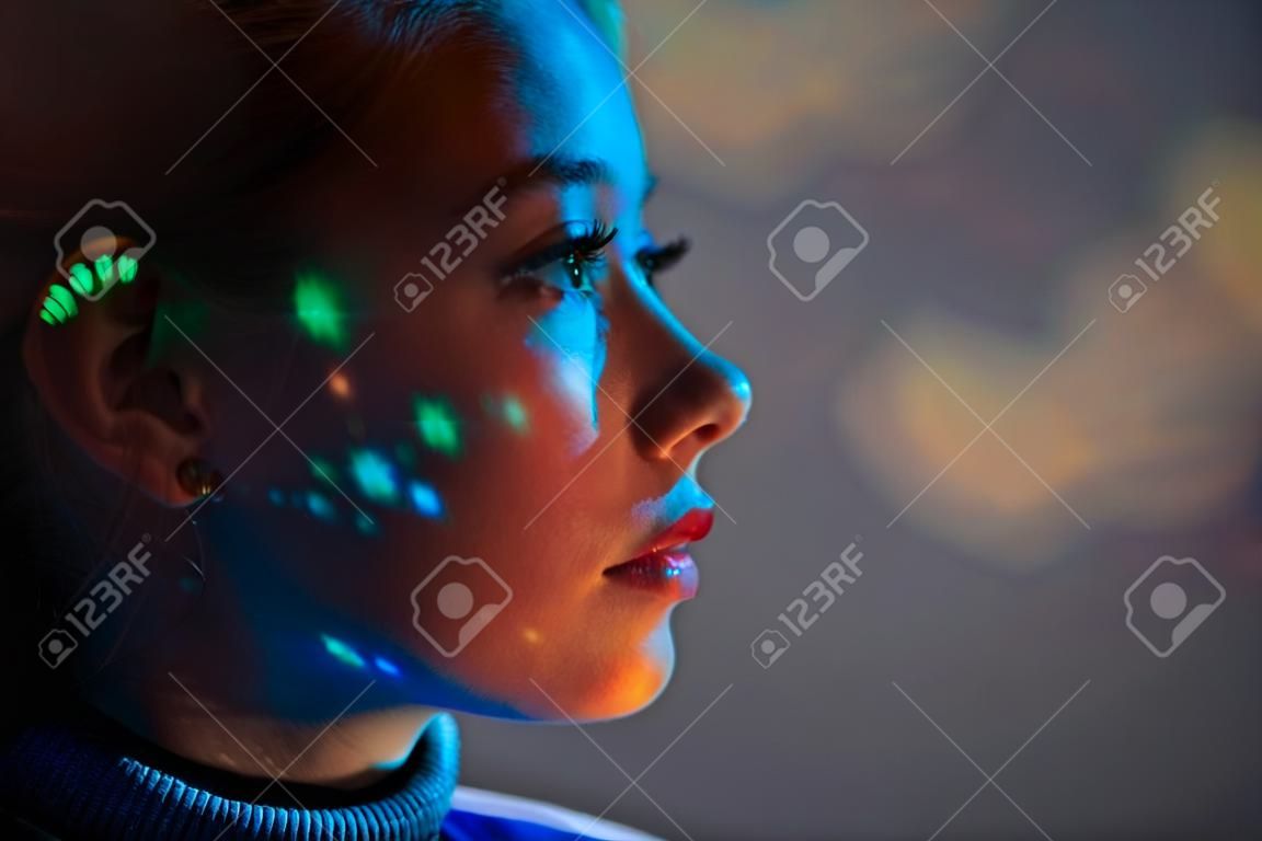 Close up portrait of beautiful tender girl looks into the distance with colored lights on your face against the background of outer space. Space concept.