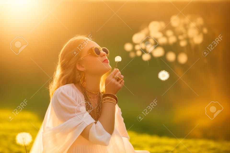 Portrait of a girl in boho style clothes and accessories joyfully blowing on dandelions in nature in warm rays of the setting sun. Bohemian, modern hippie style. Summer mood. Copy space.