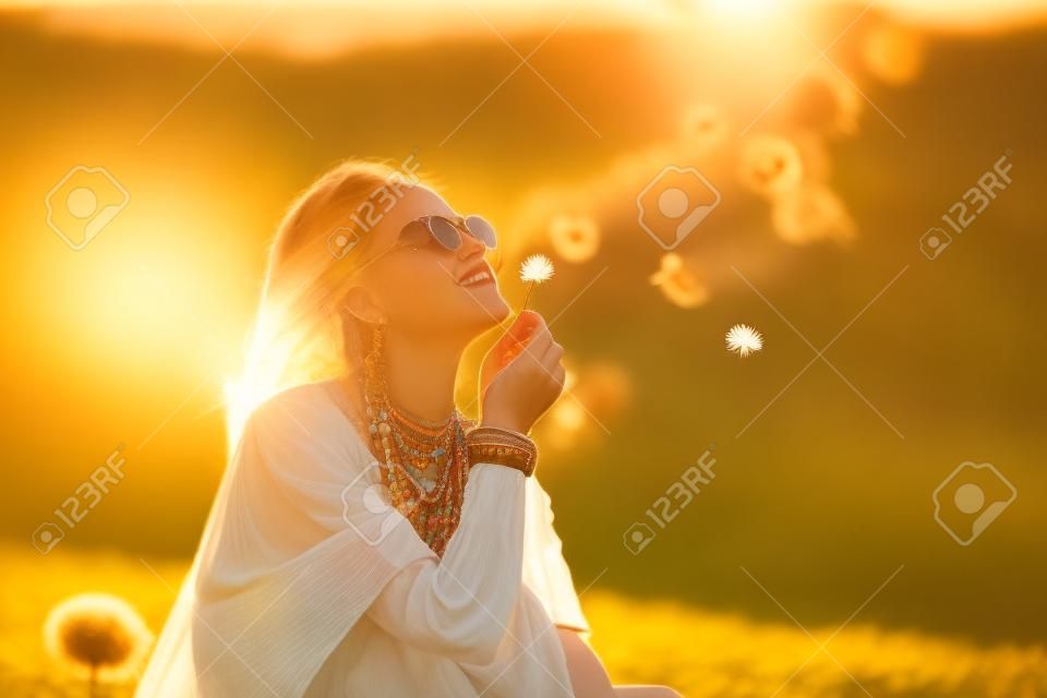 Portrait of a girl in boho style clothes and accessories joyfully blowing on dandelions in nature in warm rays of the setting sun. Bohemian, modern hippie style. Summer mood. Copy space.