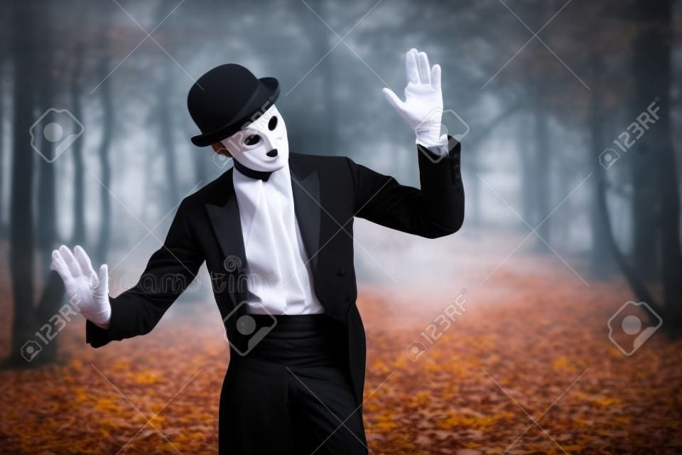 Portret tajemniczego dżentelmena w białej karnawałowej masce, meloniku i czarnym fraku stojącego w starym jesiennym parku. mim, karnawał. Halloween. zabytkowy styl.
