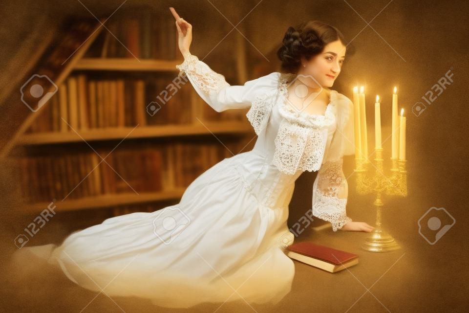 흰색 레이스 드레스를 입은 귀족 소녀가 어두운 빈티지 도서관에 앉아 읽을 책을 고르고 있습니다. 빅토리아 스타일. 역사 신비 소설.