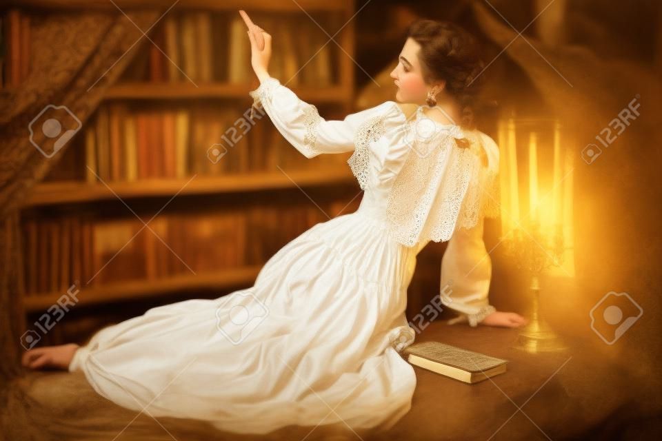 Une fille aristocratique vêtue d'une robe en dentelle blanche est assise dans une bibliothèque sombre et vintage en choisissant un livre à lire. style victorien. roman mystique historique.