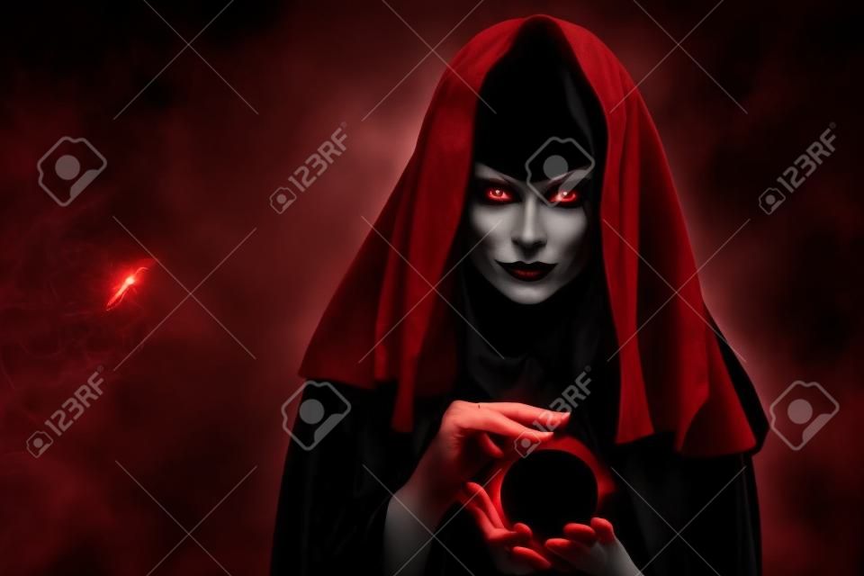 Magie noire. une sorcière noire envoûtante dans une cape rouge avec une capuche évoque avec une boule de cristal dans ses mains et regarde loin méchamment. contes magiques. halloween. espace de copie.