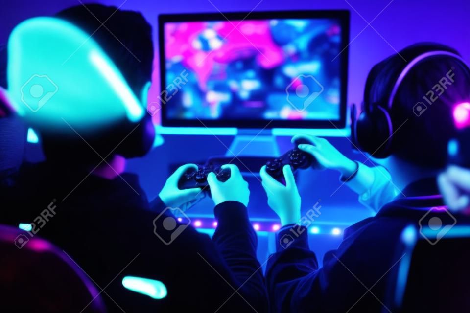 Friends playing video game at home. Gamers holding gamepads sitting at front of screen. Streamers girl and boy playing online in dark room lit by neon lights. Competition and having fun