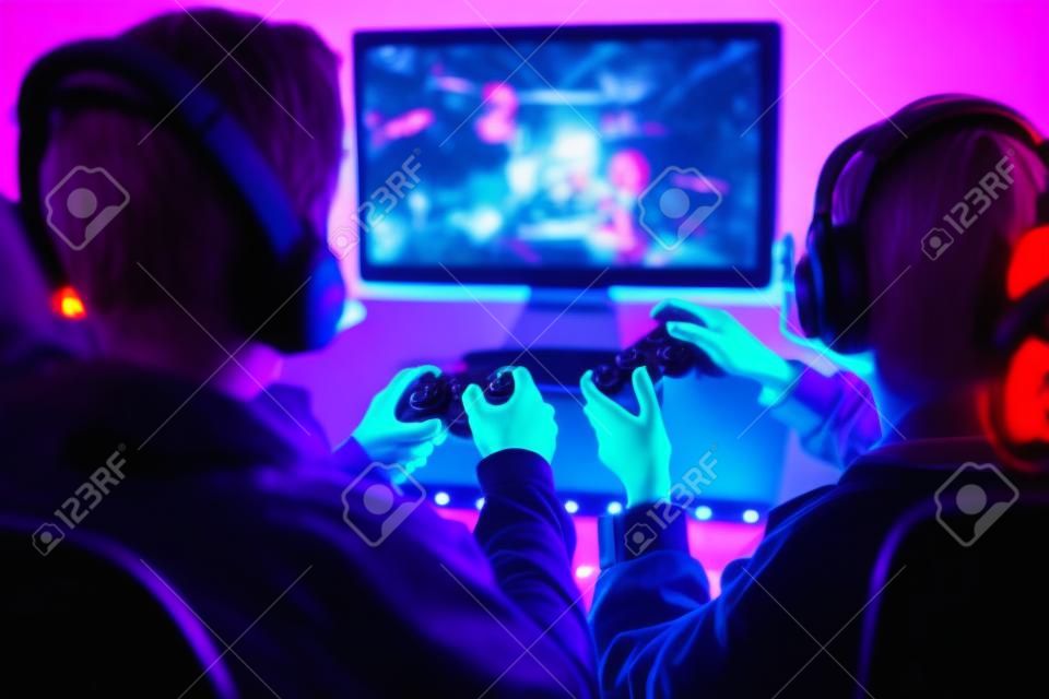Friends playing video game at home. Gamers holding gamepads sitting at front of screen. Streamers girl and boy playing online in dark room lit by neon lights. Competition and having fun