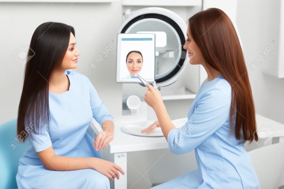 美医生给信息的女人对她的面部皮肤美容的女性美容院的人脸分析的高分辨率处理谈条件的医疗咨询特写