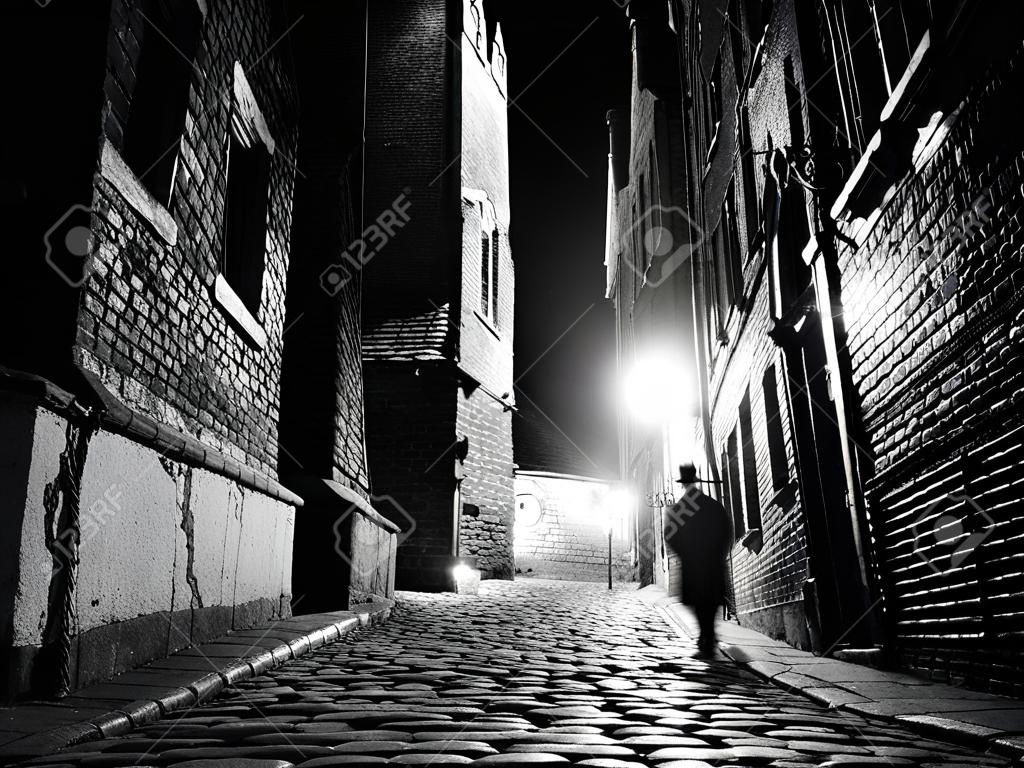 Illuminated cobbled street with light reflections on cobblestones in old historical city by night. Dark blurred silhouette of person evokes Jack the Ripper. Black and white image.