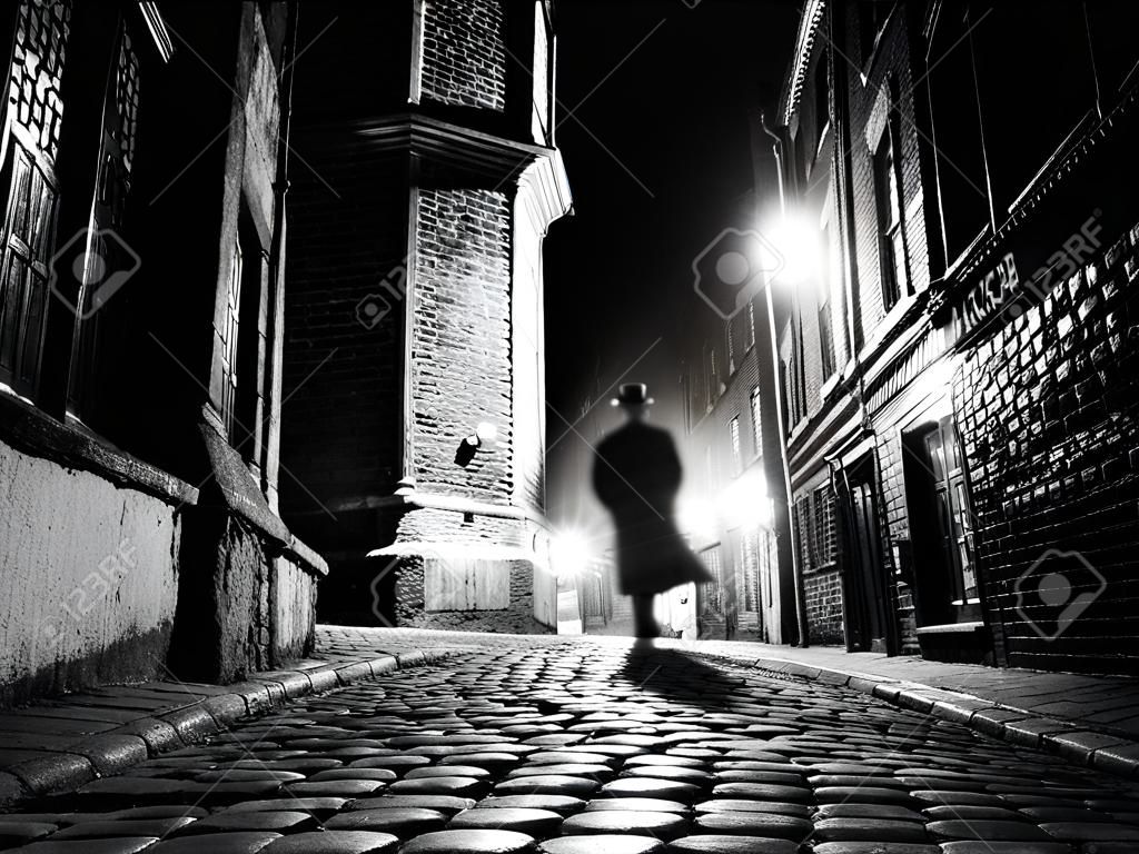 Iluminado calle empedrada con reflejos de luz en adoquines en la ciudad vieja histórica por la noche. Silueta borrosa Oscuro de persona evoca Jack el Destripador. Imagen blanco y negro.