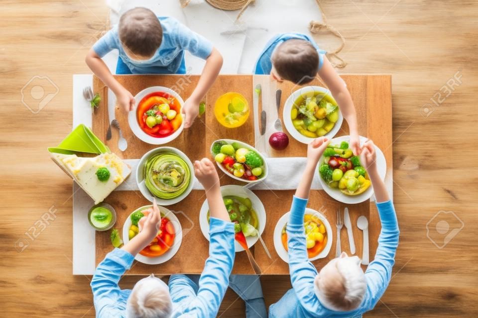 Draufsicht auf Eltern und Kinder, die am Küchentisch sitzen und gemeinsam lecker zu Mittag essen: frischer Gemüsesalat, Obst, Käse und hausgemachter Kuchen