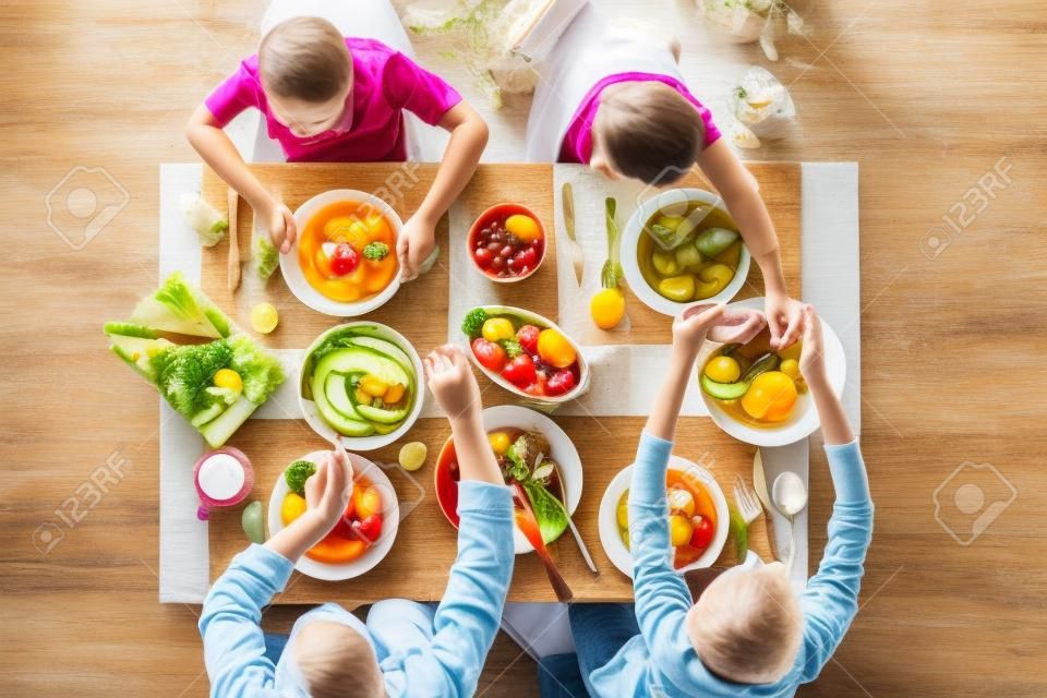 Draufsicht auf Eltern und Kinder, die am Küchentisch sitzen und gemeinsam lecker zu Mittag essen: frischer Gemüsesalat, Obst, Käse und hausgemachter Kuchen