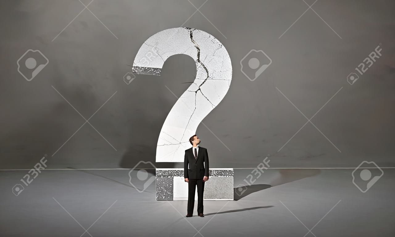 Una pequeña persona de negocios elegante en traje de pie con la espalda delante de un gran signo de interrogación en el concepto de espacio abierto