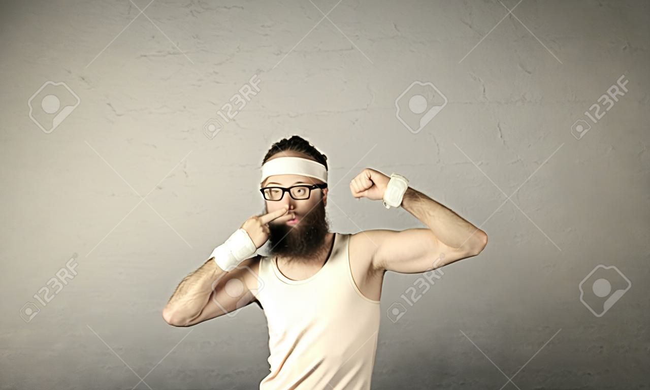A young man with beard, headstrap and glasses posing in front of blank grey wall background, imagining he has big muscles