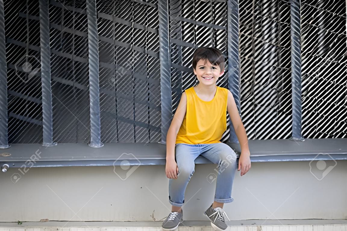 Widok z przodu uśmiechniętego chłopca siedzącego na metalowym ogrodzeniu, patrzącego na kamerę w jasny dzień