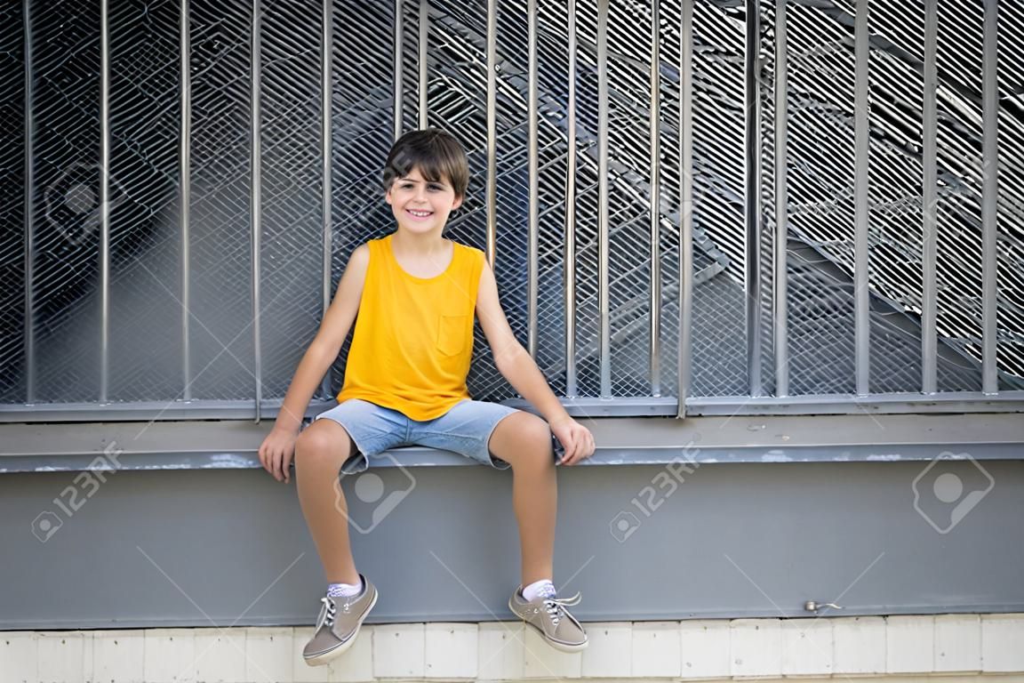 Widok z przodu uśmiechniętego chłopca siedzącego na metalowym ogrodzeniu, patrzącego na kamerę w jasny dzień