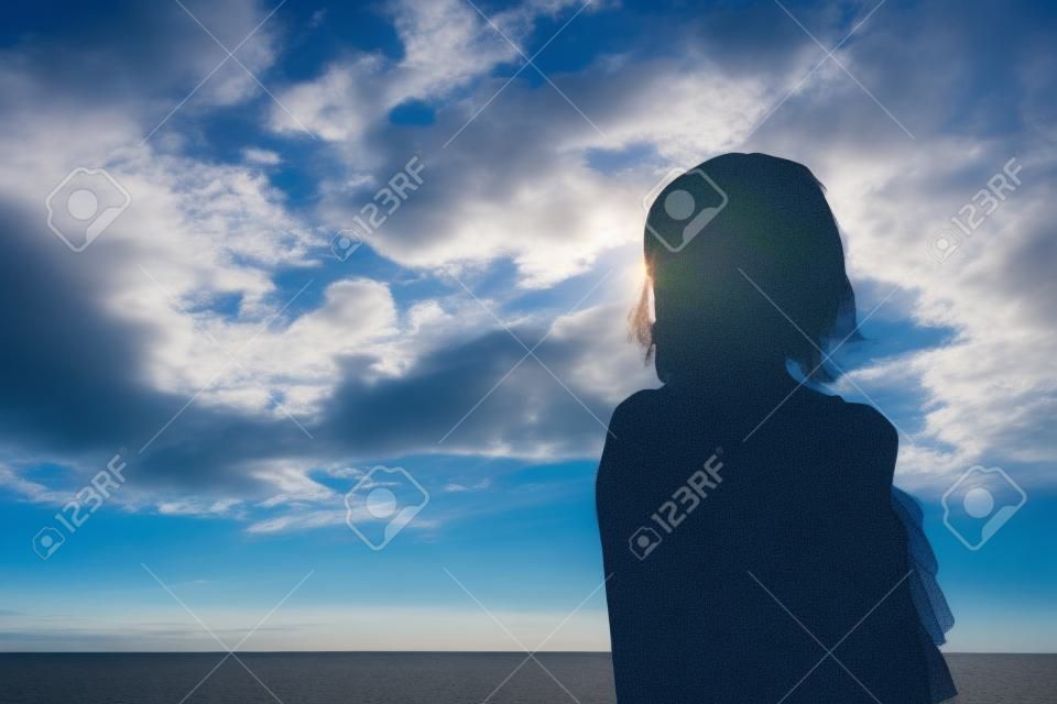 Sylwetka kobiety na tle pochmurnego, błękitnego nieba rano na plaży, gdy wschodzi słońce, stojąc pewnie i patrząc na cień