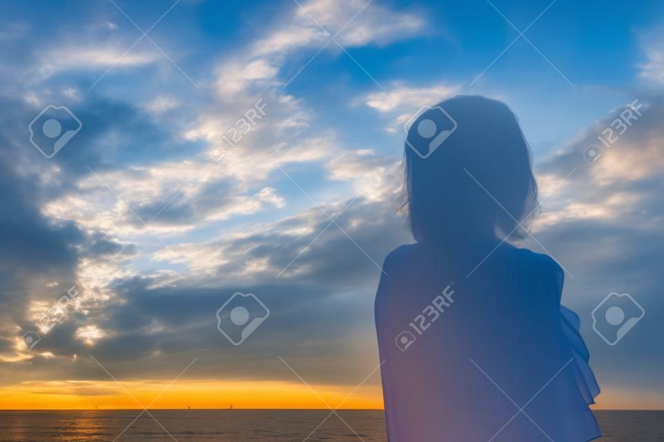Silueta de dama contra el cielo azul nublado por la mañana en la playa cuando sale el sol, de pie confiada con la sombra mirando