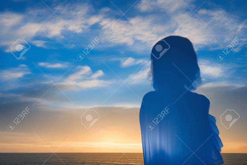 Damensilhouette gegen bewölkten blauen Himmel morgens am Strand bei Sonnenaufgang, selbstbewusst mit Schattenblick