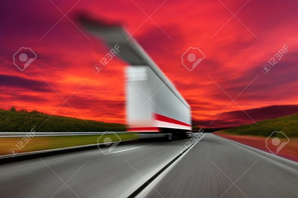 흐리게. 대형 트럭이 고속도로를 따라 고속으로 주행하고 있습니다. 밝은 붉은 구름이 있는 하늘. 화물 인도.