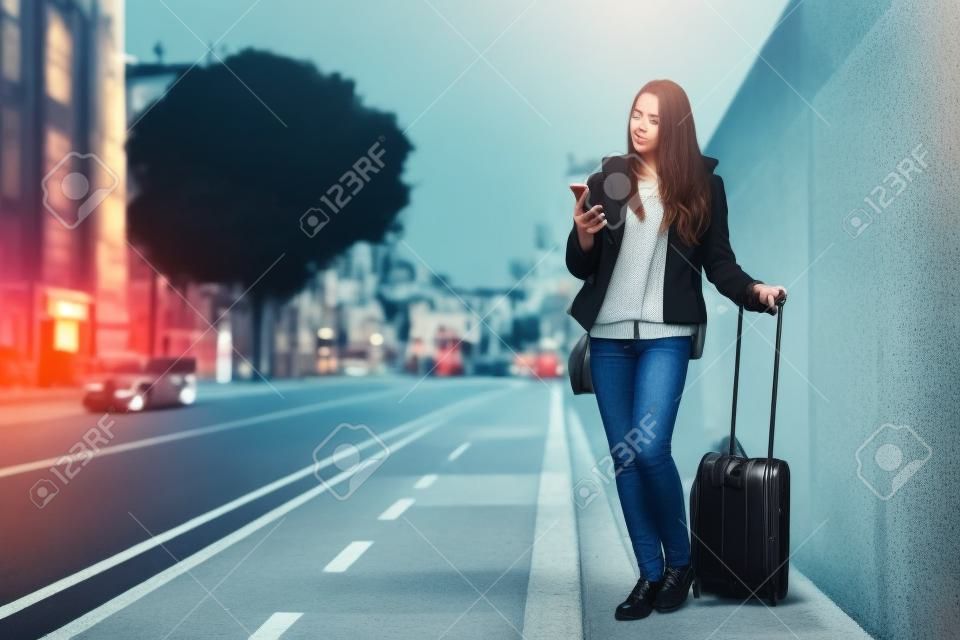 Młoda kobieta z walizką czeka na taksówkę na ulicy, konsultując się z telefonem, koncepcją podróży i technologii, miejscem na tekst