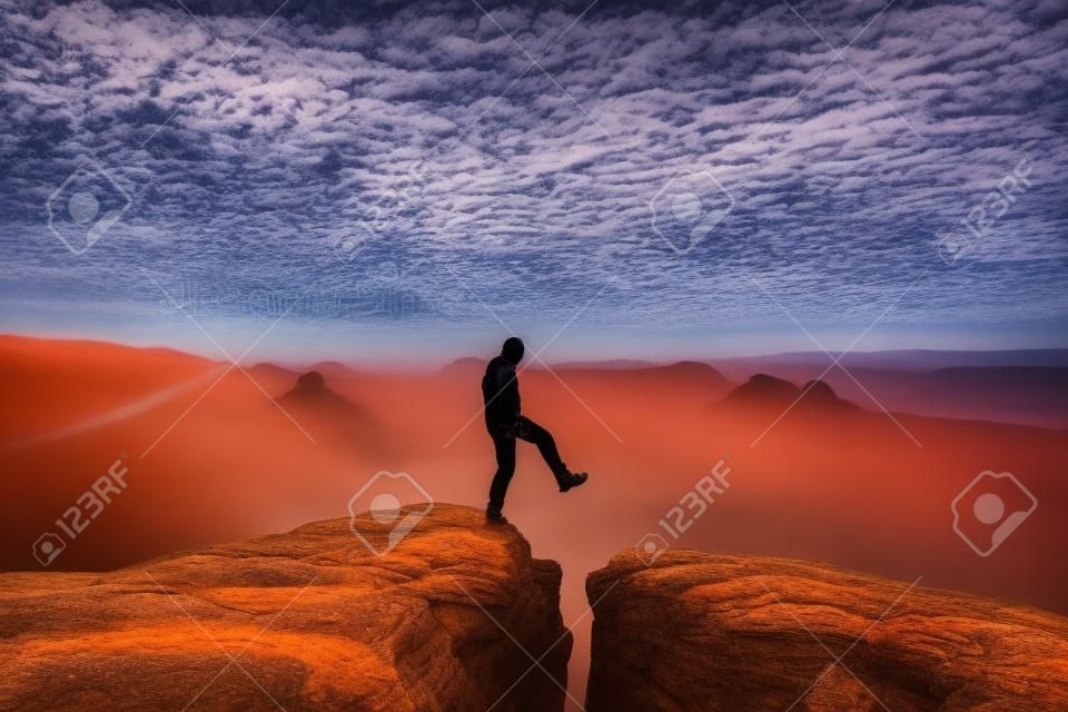 Man in black jumping between rocky empires. Dreamy daybreak in sandstone mountains, heavy orange mist in deep valley.