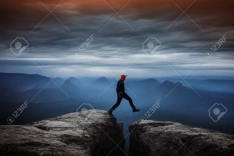 Caminhante louco em preto está pulando entre picos rochosos. Homem feliz. Maravilhoso amanhecer em montanhas rochosas, névoa laranja pesada no vale profundo. Milagre da natureza