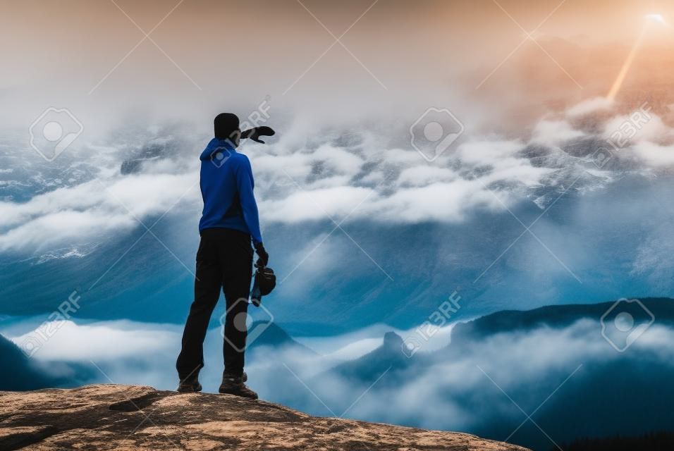 Male hiker is stand on edge and enjoying dramatic overlook of misty landscape. The Sun is hidden in thick mist and change colors.