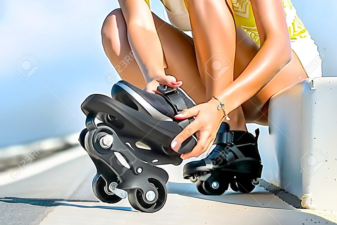 Woman lacing up rollers on the asphalt road near the sea in summer. Close up view focused on hands and rollers