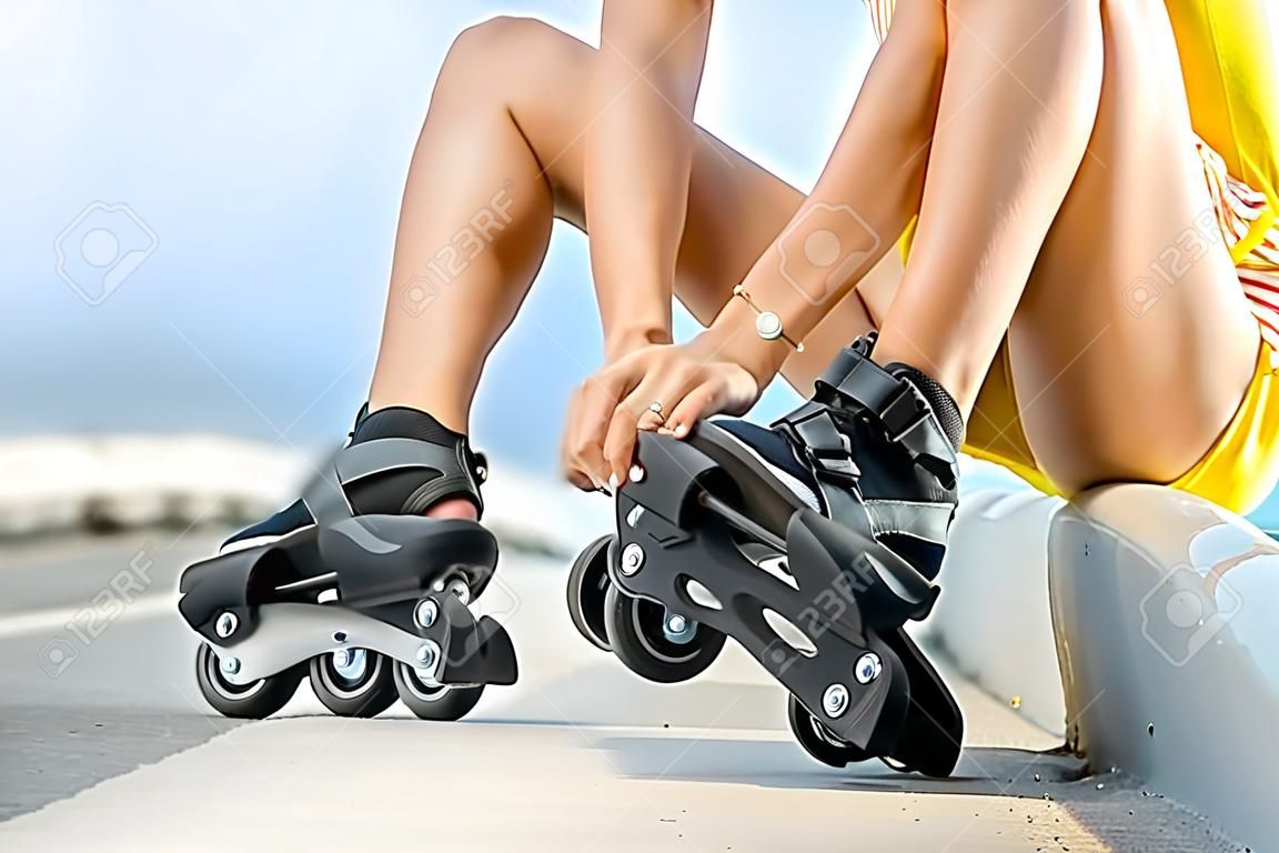 Woman lacing up rollers on the asphalt road near the sea in summer. Close up view focused on hands and rollers