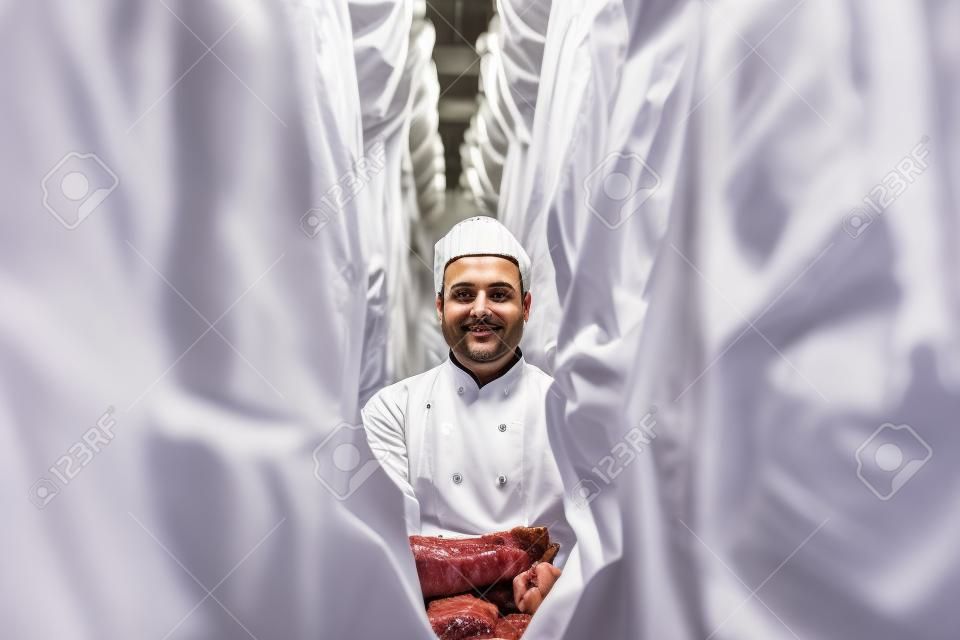 Portrait of a handsome butcher in white uniform with pork carcasses at the meat manufacturing