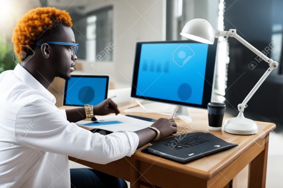 Junger afrikaner, der mit analitycs arbeitet, sitzt am schönen arbeitsplatz mit computer, laptop und dokumenten