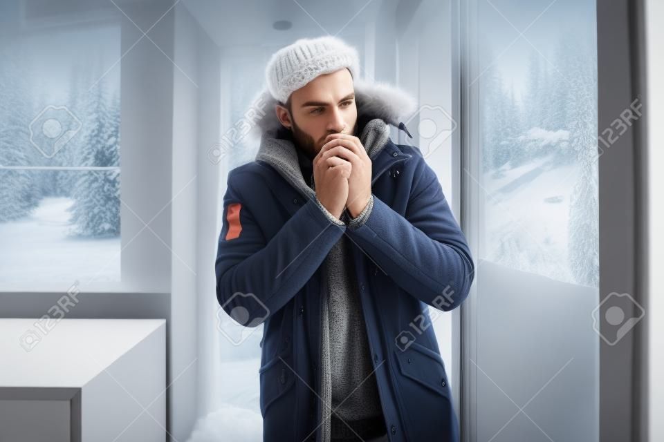 Hombre vestido con ropa de invierno sintiendo frío de pie cerca de la ventana en casa sin calefacción