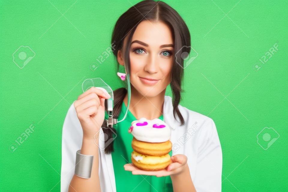 Ritratto di un nutrizionista donna triste in abito medico con ciambelle e metro a nastro sullo sfondo verde. Mangiare malsano e concetto di adiposità