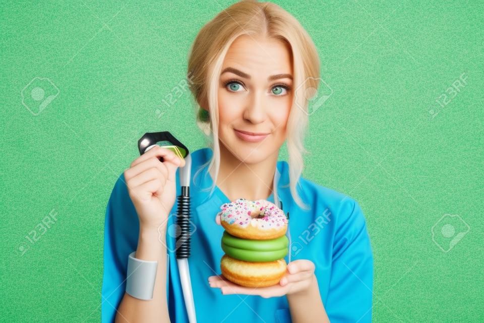 Ritratto di un nutrizionista donna triste in abito medico con ciambelle e metro a nastro sullo sfondo verde. Mangiare malsano e concetto di adiposità