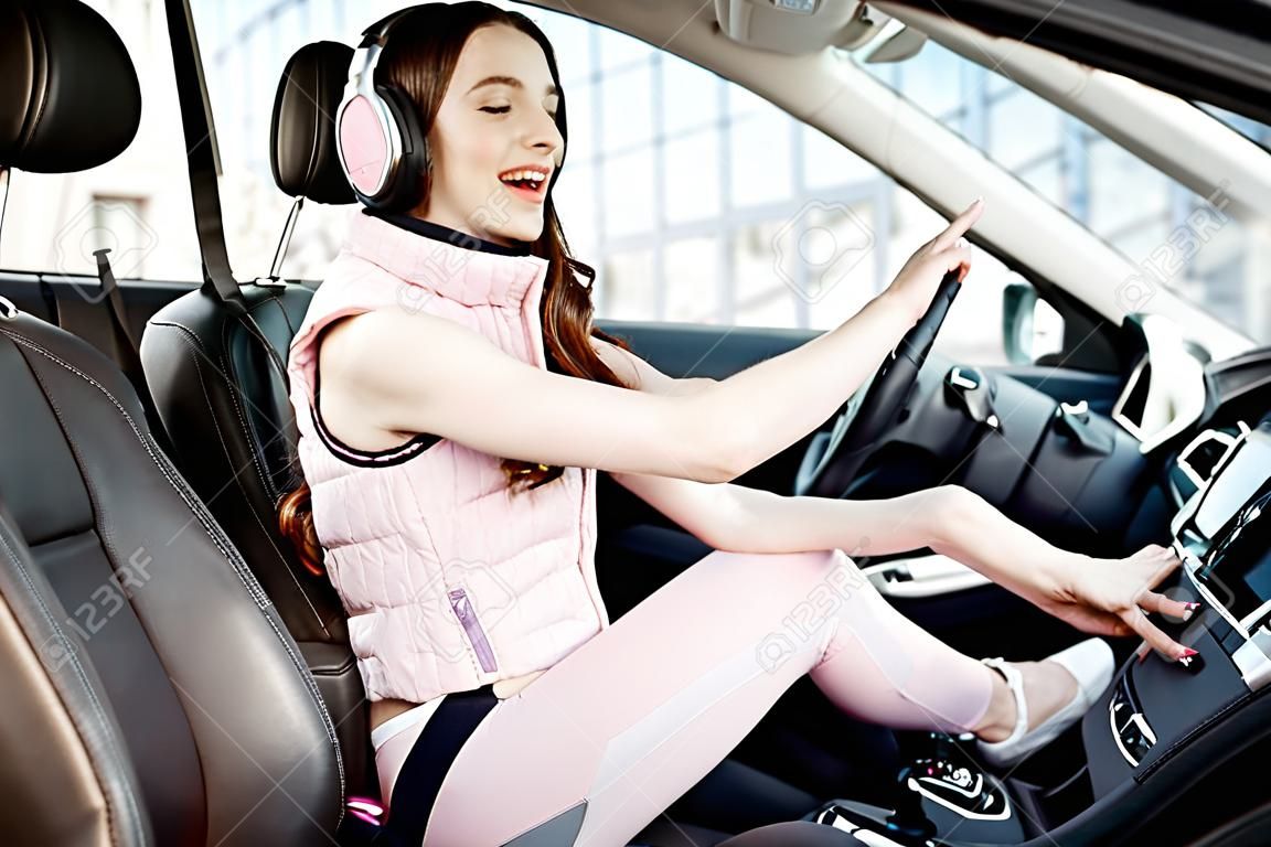 Giovane e allegra donna in abiti sportivi rosa che ascolta la musica mentre guida un'auto di lusso in città