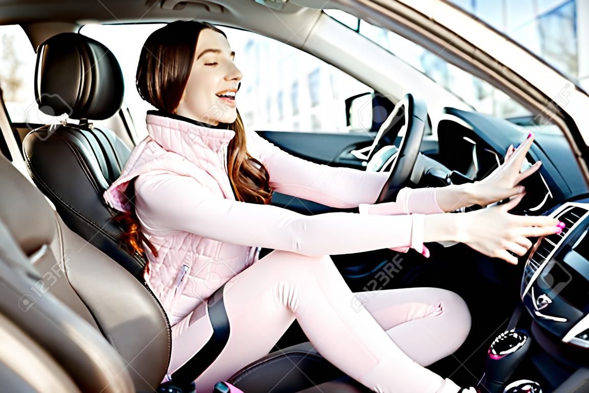 Young and cheerful woman in pink sportswear listening to the music while driving luxury car in the city