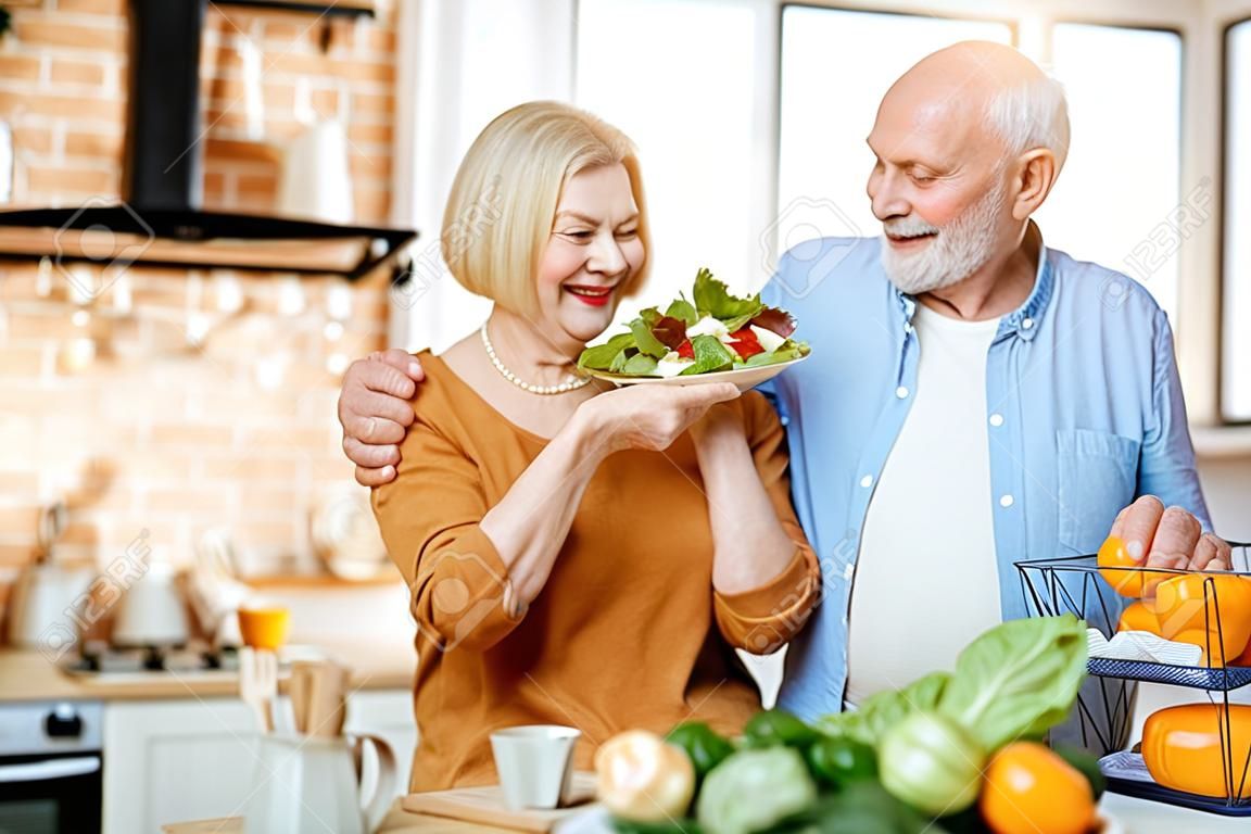 Ritratto di una coppia senior allegra con insalata e cibo sano in cucina a casa. Concetto di alimentazione sana in età avanzata