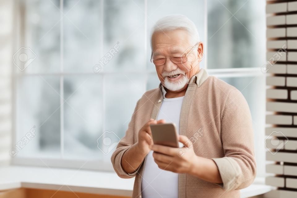 Cheerful senior grandfather using phone standing near the window at home