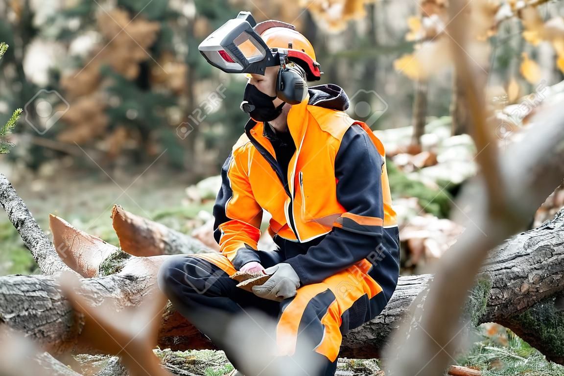 Retrato de um madeireiro profissional em vestuário de trabalho de proteção sentado com uma motosserra na árvore derrubada, descansando após o trabalho duro na floresta