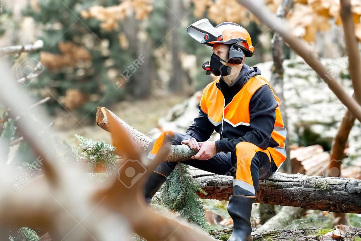 Retrato de um madeireiro profissional em vestuário de trabalho de proteção sentado com uma motosserra na árvore derrubada, descansando após o trabalho duro na floresta