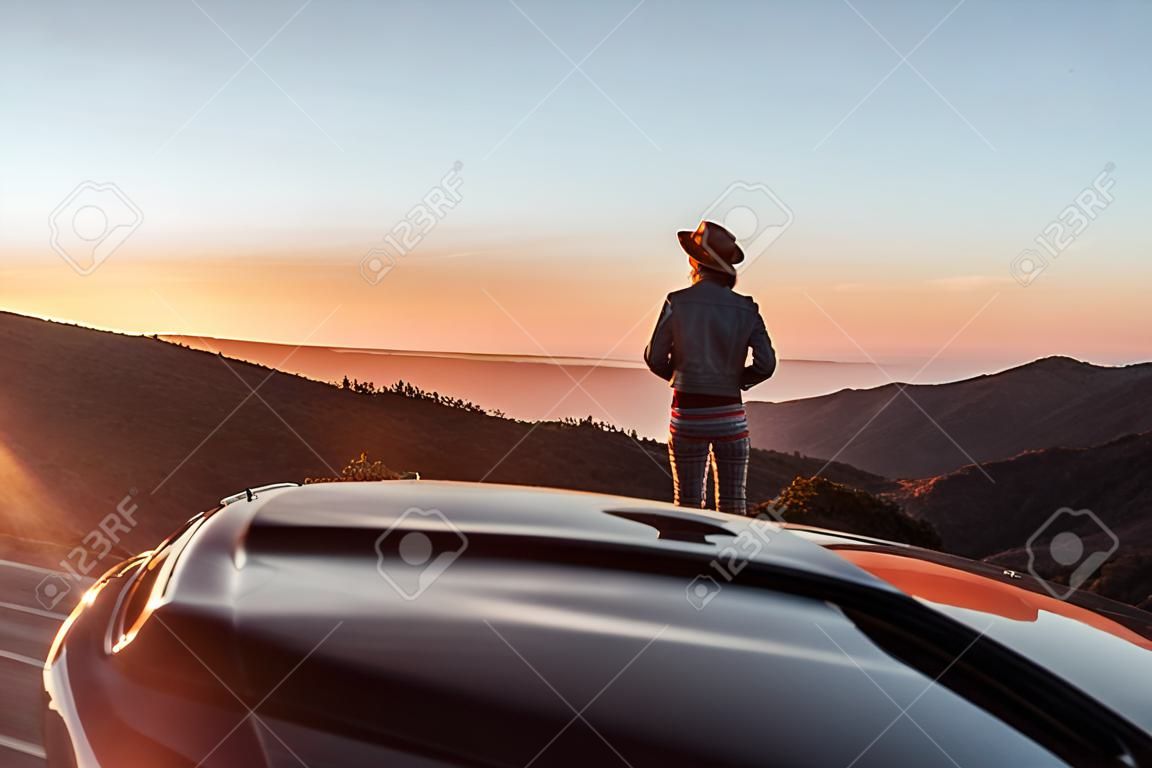 Widok na krajobraz na poboczu drogi nad chmurami z kobietą podziwiającą piękny zachód słońca podczas podróży kabrioletem sportowym