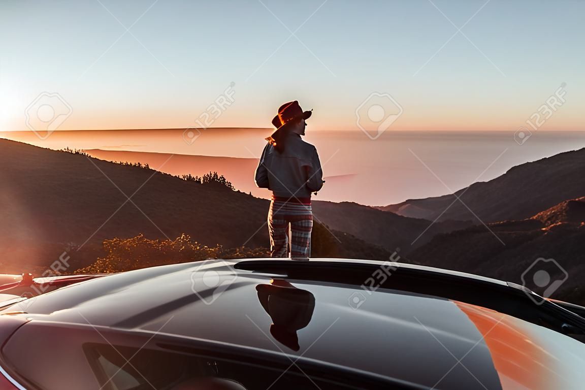 Landschaftsansicht am Straßenrand über den Wolken mit einer Frau, die einen wunderschönen Sonnenuntergang genießt, während sie mit dem Cabrio-Sportwagen unterwegs ist