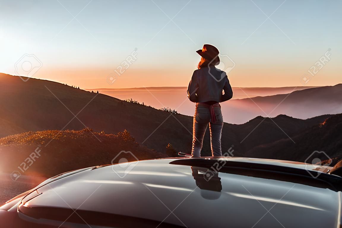 コンバーチブルスポーツカーで旅行中に美しい夕日を楽しむ女性と雲の上の道端の風景ビュー