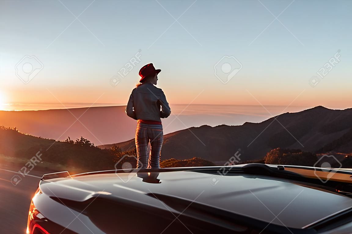 Vista da paisagem na beira da estrada acima das nuvens com mulher desfrutando de belo pôr do sol enquanto viaja no carro esportivo conversível