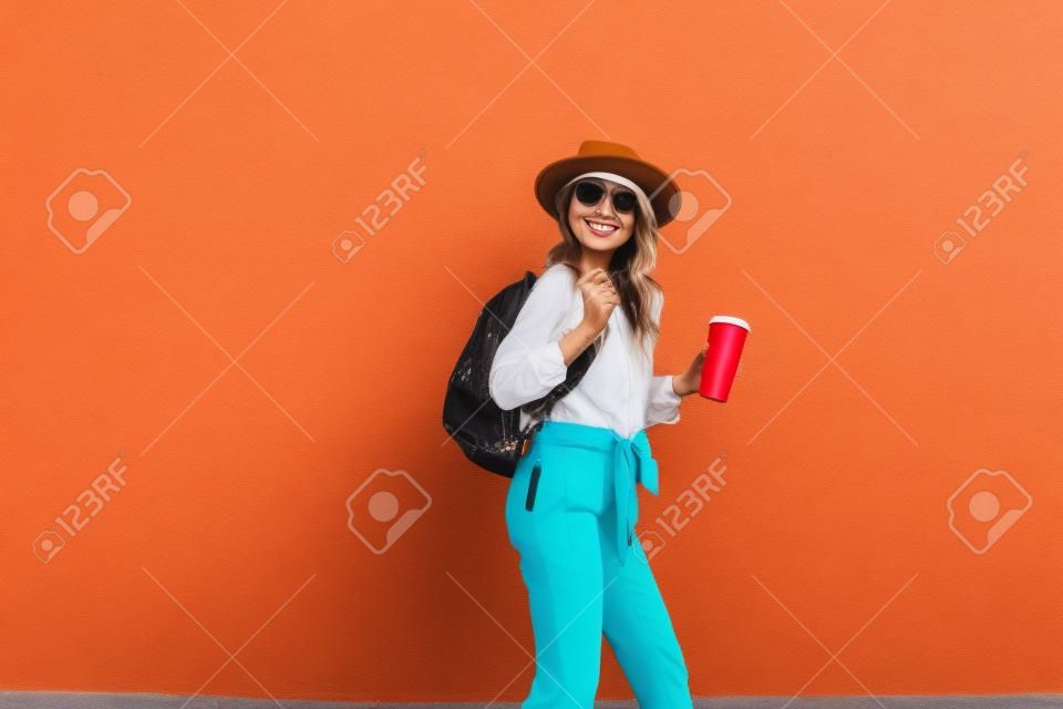 Portret aktywnej stylowej kobiety ubranej w jasną koszulę z kapeluszem i filiżanką kawy na tle czerwonej ściany na zewnątrz. Beztroski styl życia, kawa i koncepcja mody damskiej