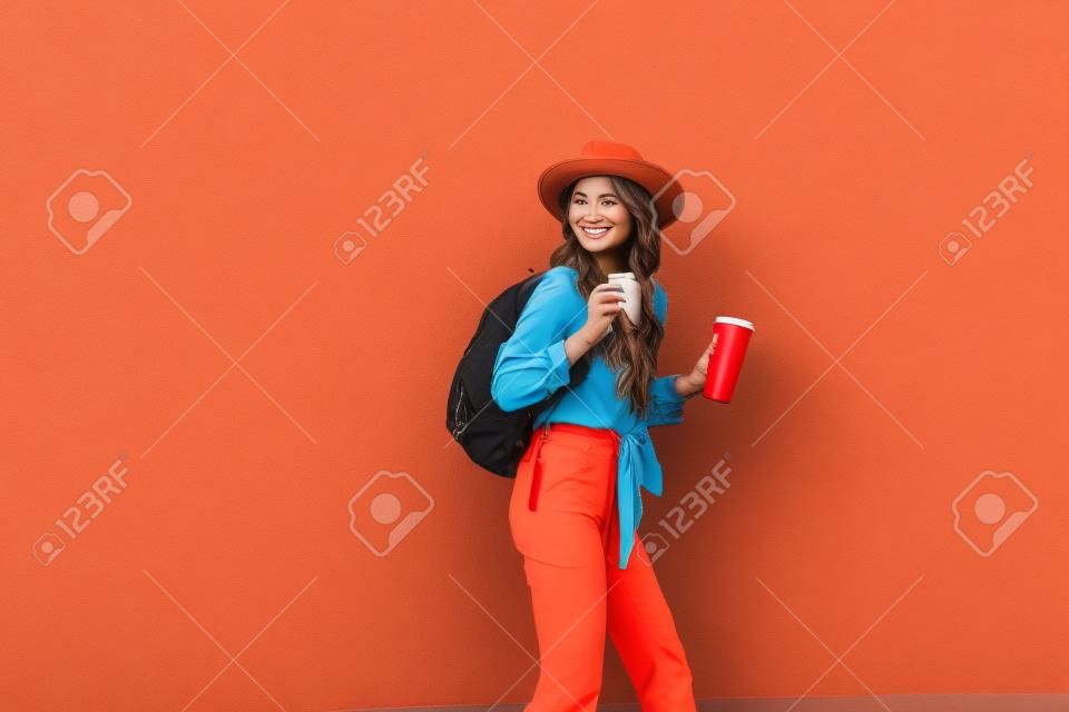 Portret aktywnej stylowej kobiety ubranej w jasną koszulę z kapeluszem i filiżanką kawy na tle czerwonej ściany na zewnątrz. Beztroski styl życia, kawa i koncepcja mody damskiej