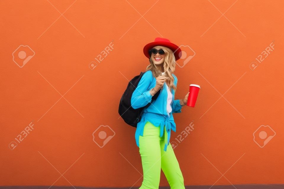 Portrait of an active stylish woman dressed in bright shirt with hat and coffee cup on the red wall background outdoors. Carefree lifestyle, coffee and womens fashion concept